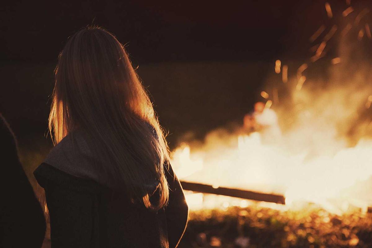 Person standing by a large bonfire, with warm glow illuminating their long hair in a cozy outdoor setting.