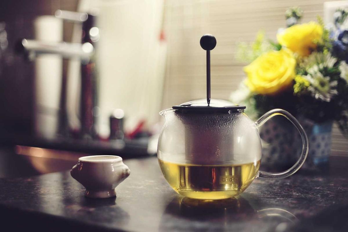 A glass teapot with a brewing infuser stands next to a small white cup on a kitchen countertop.