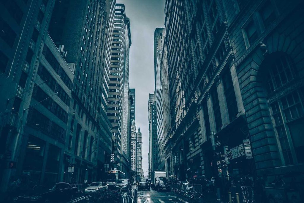Cityscape view of bustling urban street with tall buildings under a cloudy sky, cars and pedestrians.