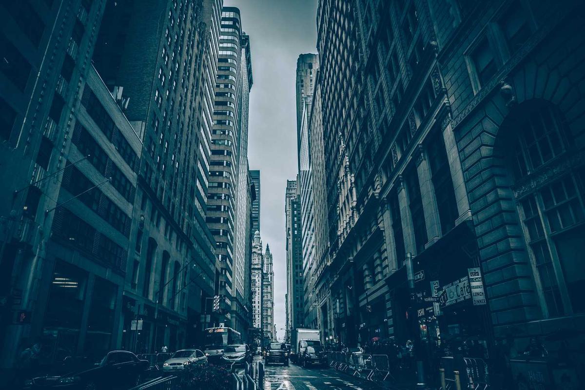 Cityscape view of bustling urban street with tall buildings under a cloudy sky, cars and pedestrians.