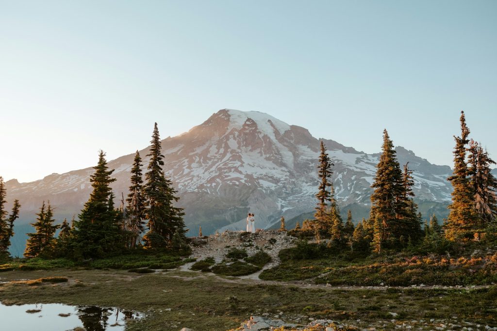 Couple embracing in front of a majestic mountain at sunset, surrounded by evergreen trees and alpine scenery.