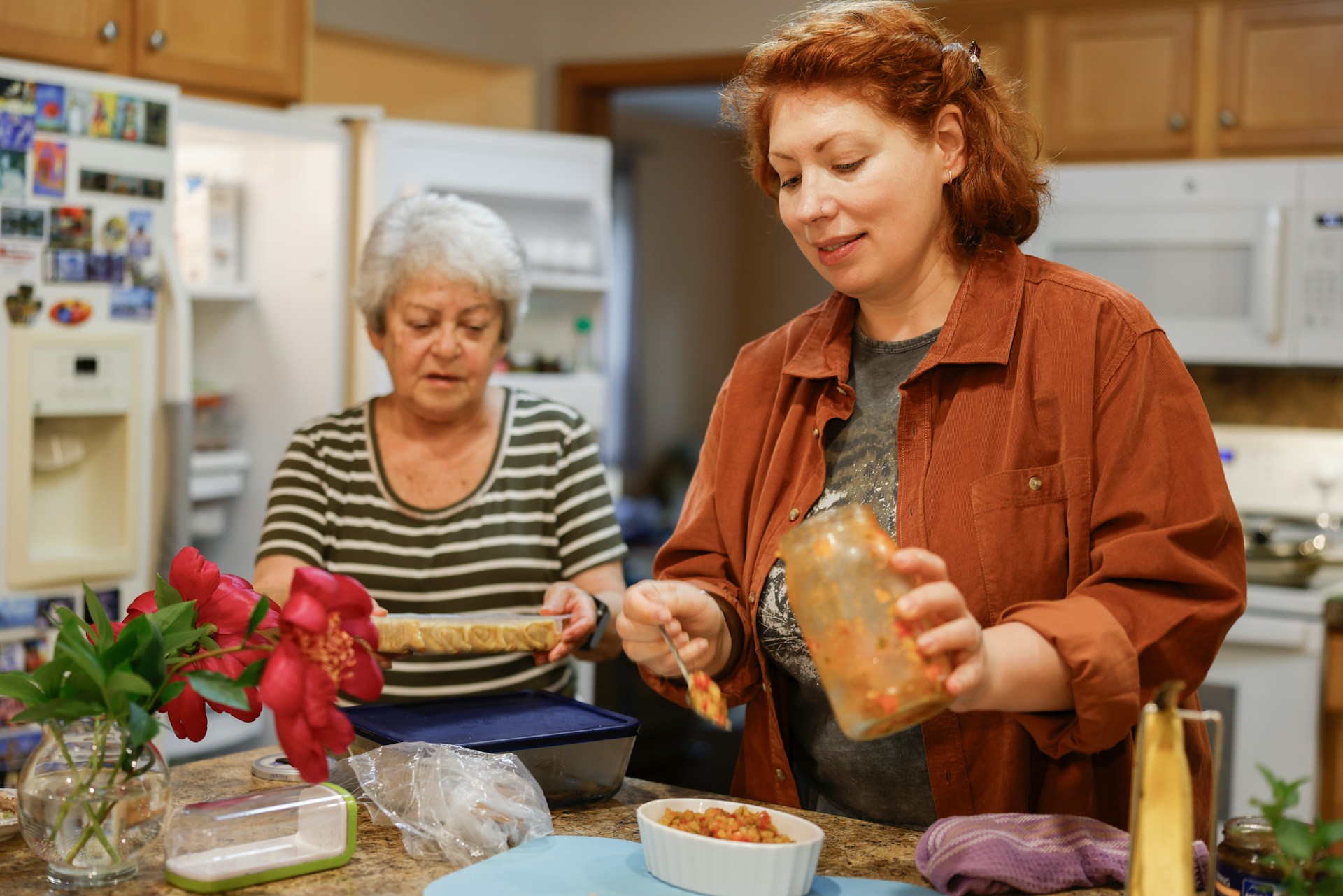 Two women cooking together in a cozy kitchen, surrounded by ingredients and fresh flowers.