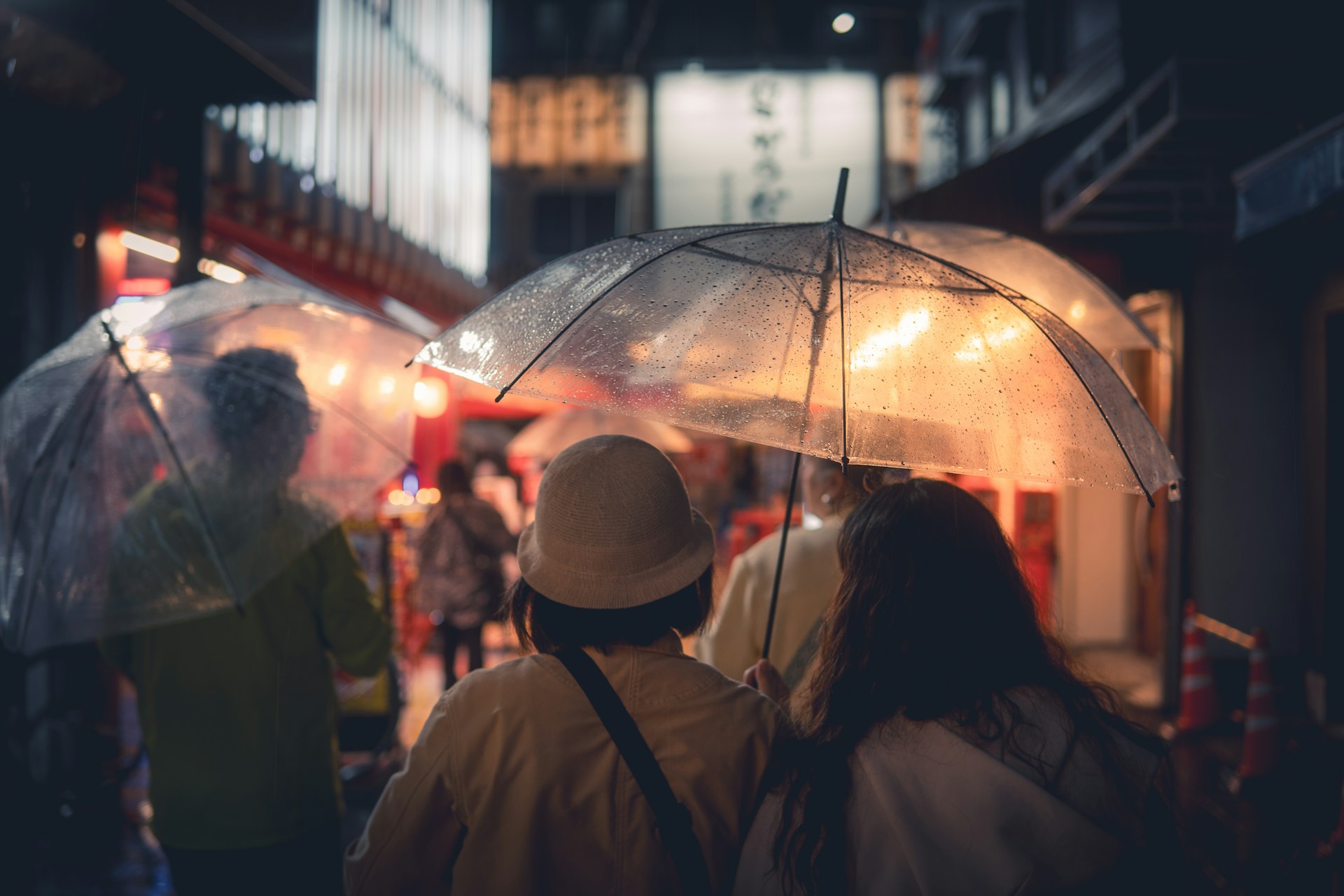 Two people holding umbrellas walk through a rainy street, illuminated by soft lights and vibrant colors at night.